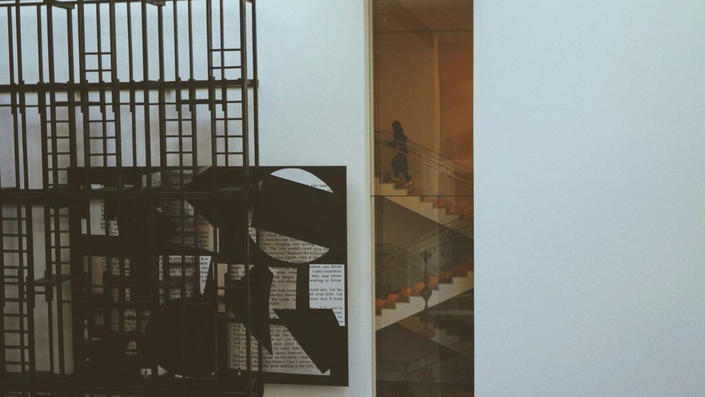 A visitor climbing a glass-walled staircase inside MoMA, seen past a black metal sculpture