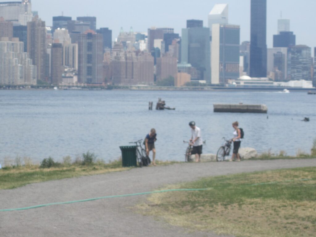 Three people with bicycles on the shoreline of the Williamsburg state park, with Midtown Manhattan across the East River