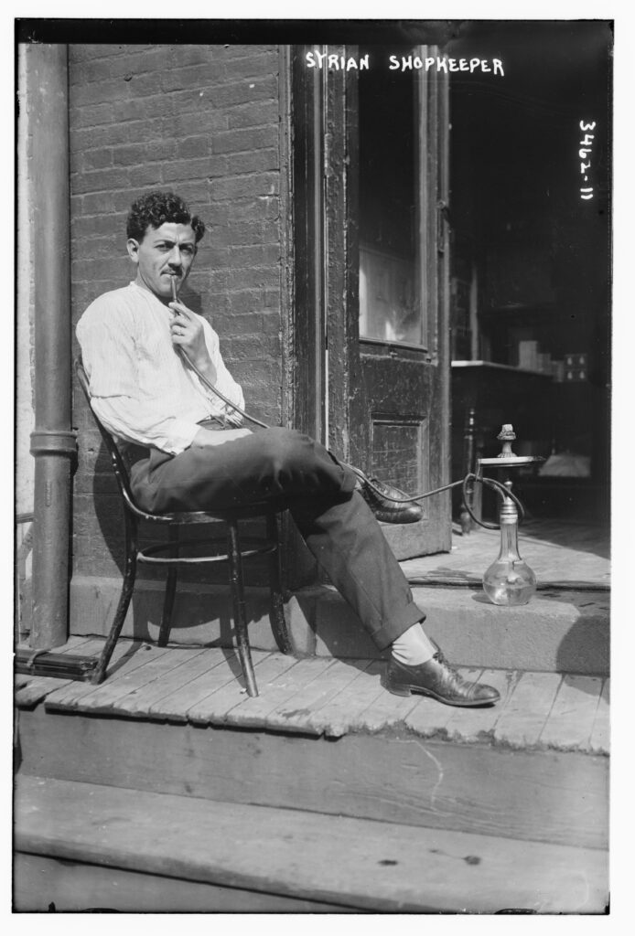 Early 1900s photograph captioned Syrian shopkeeper: a man on a chair outside his shop door with a water pipe beside him